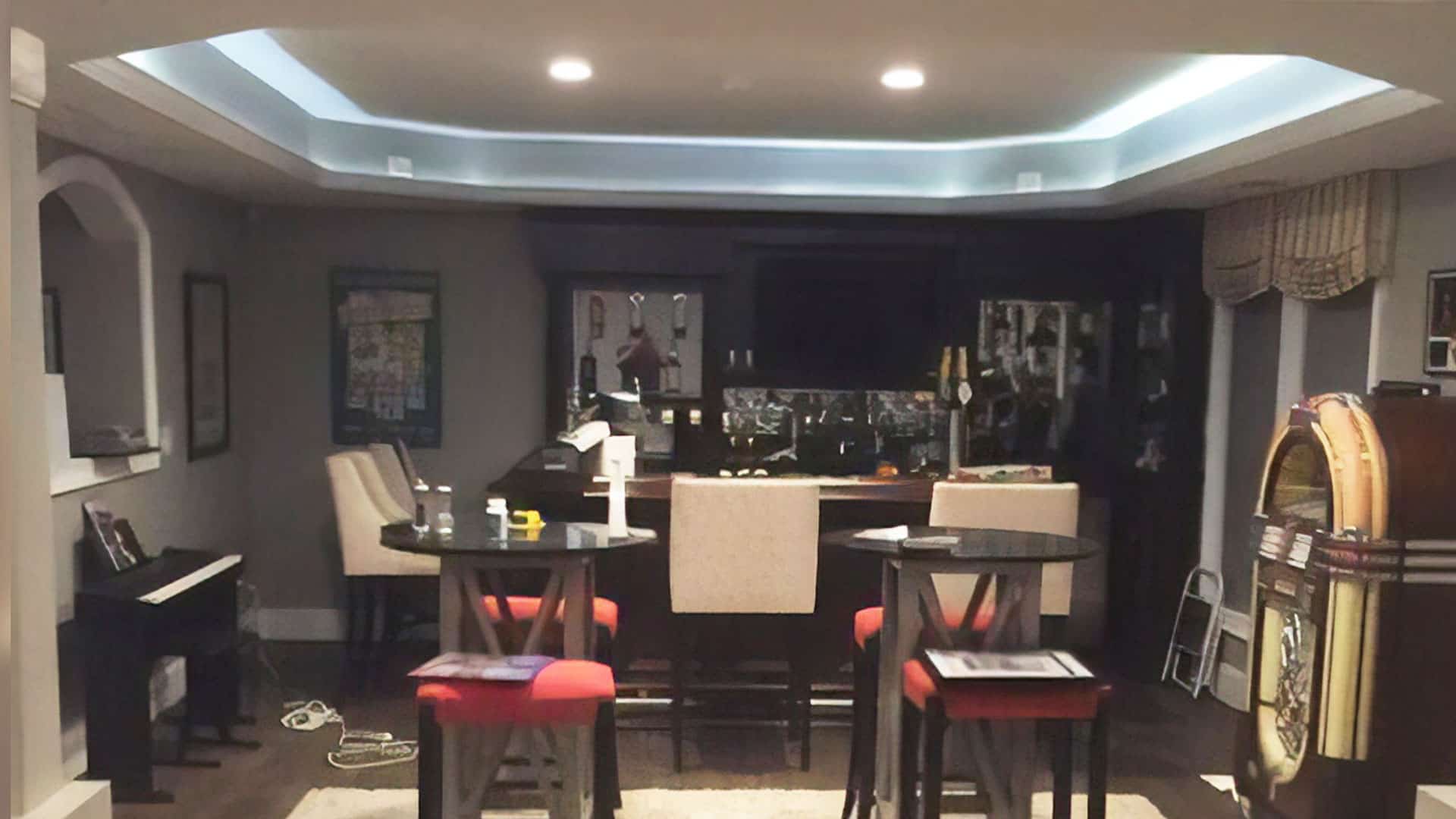 A home basement bar with high-top tables and red-cushioned chairs, a jukebox, a piano, and various decorations on the walls, under dim lighting.