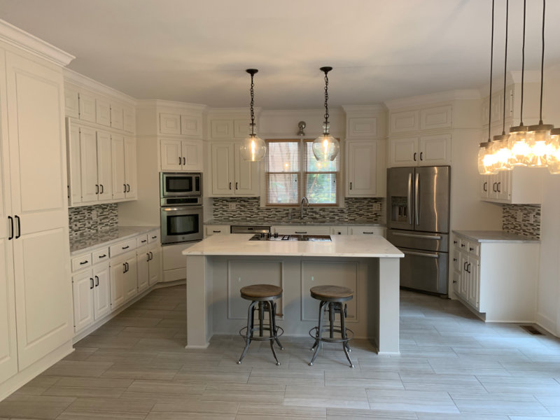 A modern white kitchen with an island, two bar stools, hanging light fixtures, stainless steel appliances, and tile flooring.