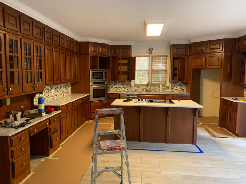 A kitchen under renovation with wooden cabinets, a central island, a step ladder, and tools placed on countertops. The countertops and floor are partly unfinished, and a window is visible above the sink.