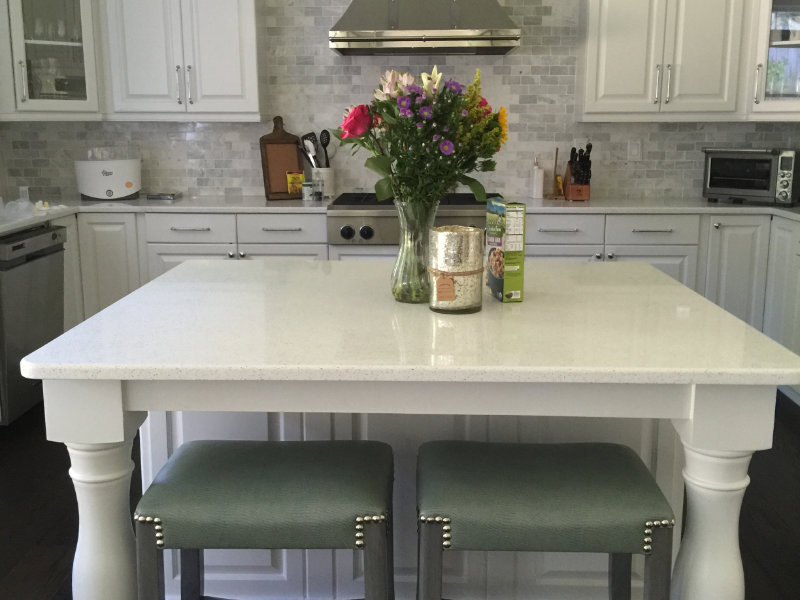 A modern kitchen features a white island with two stools, a vase of flowers, a candle, and a box of cookies. White cabinets and stainless steel appliances are in the background.