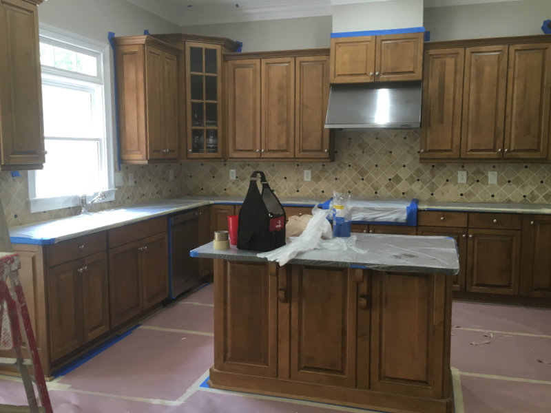 A kitchen with wooden cabinets and countertops covered for renovation. A black bag and various tools are on the island counter. The floor is protected with taped-down paper.