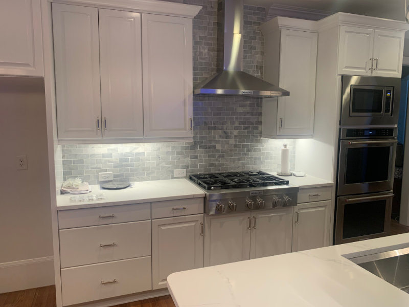 Modern kitchen with white cabinets, silver drawer handles, and a stainless steel gas stove and oven. The backsplash features light gray tiles. Bright under-cabinet lighting illuminates the countertop.