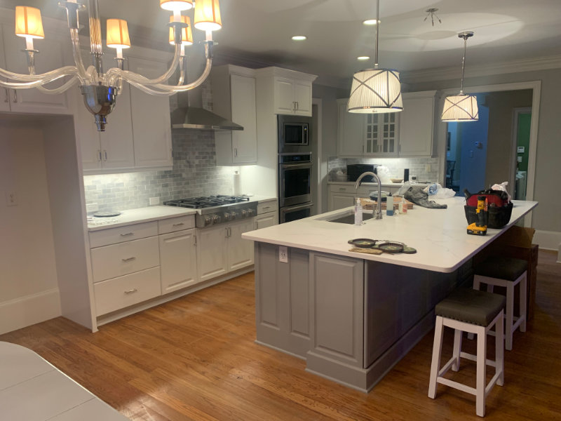 A modern kitchen with white cabinetry, a grey island, stainless steel appliances, and wooden flooring. Chandelier and hanging lights illuminate the space. Tools and supplies are on the island.