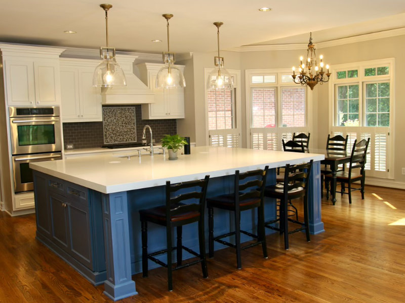 Modern kitchen with a large island featuring blue cabinetry and white countertop. Equipped with stainless steel appliances, pendant lighting, chandelier, wooden flooring, and a dining area with six chairs.