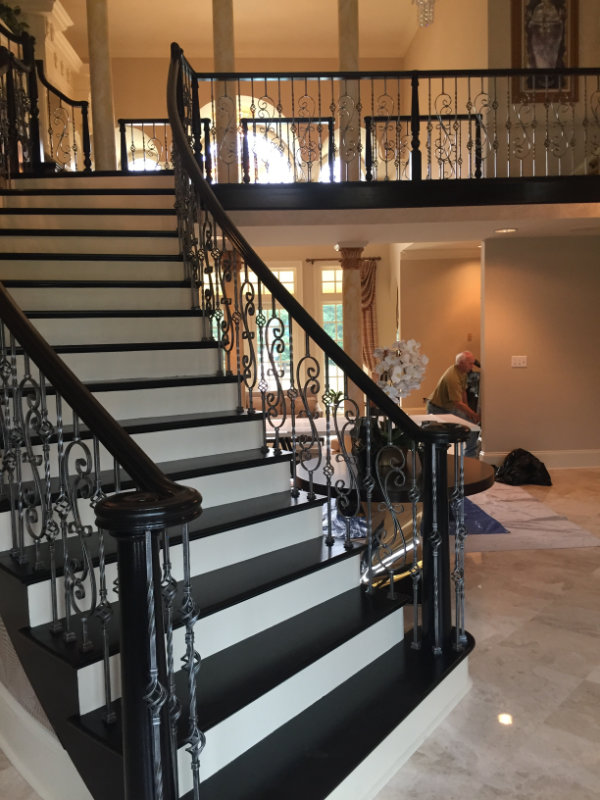 A curved staircase with ornate iron railings leads to an upper floor. The lower level has a marble floor with a person working in the background near a wall and a table with a decorative item.