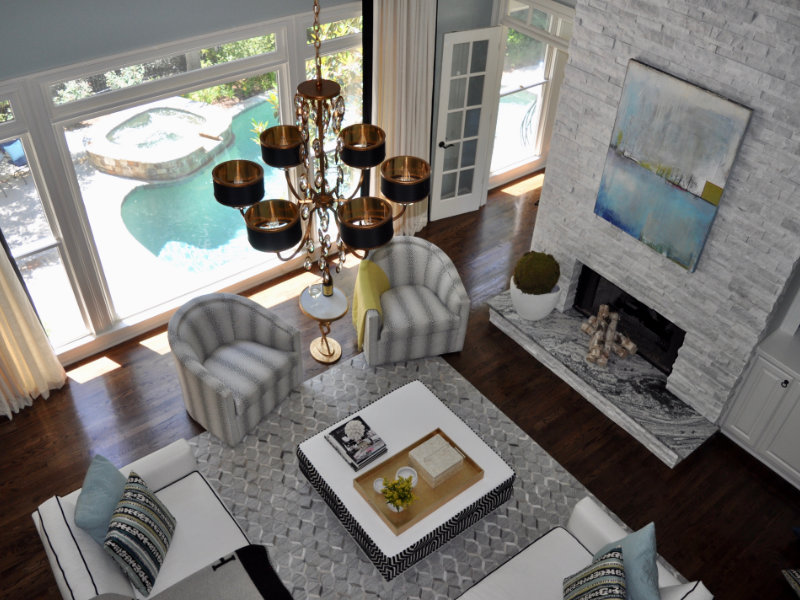 A modern living room with a large chandelier, four armchairs, a white coffee table, and a fireplace. Floor-to-ceiling windows provide a view of a pool and patio area outside.