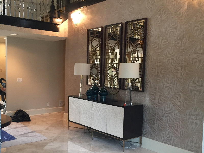 A modern living room featuring a white and dark console table, two lamps, decorative vases, and three large mirrored wall panels. The room has beige patterned wallpaper and a partial view of an upper floor.