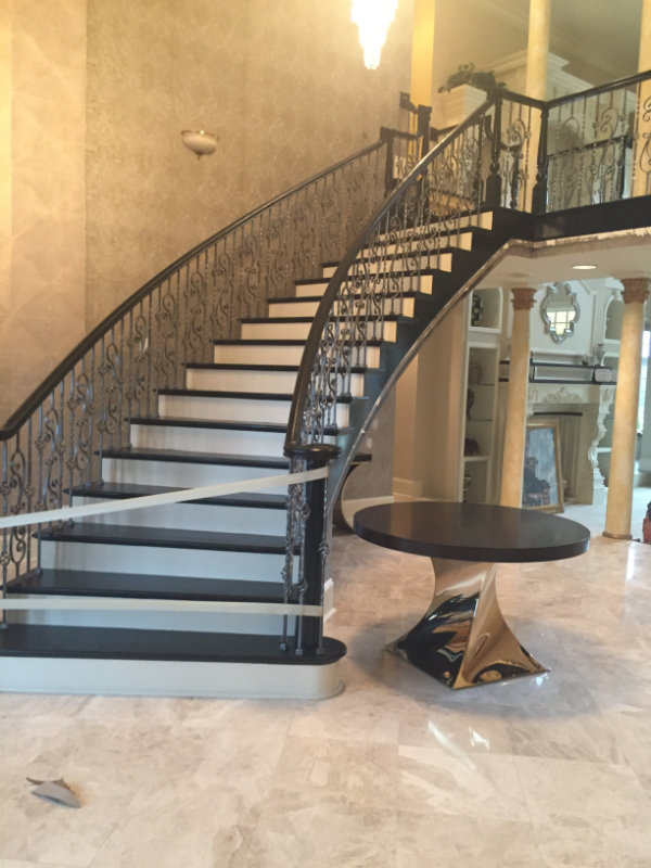 A grand curved staircase with ornate railings leads up from a marble-floored foyer, featuring a dark, round table with a reflective base at the center.