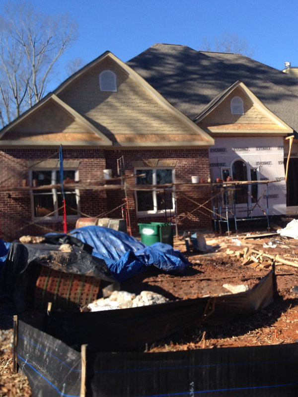 A partially constructed house with scaffolding and materials scattered in the dirt-filled front yard. The building has brick walls, and sections are covered with house wrap.