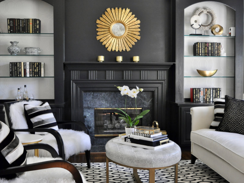 A stylish living room with a black fireplace, gold sunburst mirror, built-in shelves with books and decor, white furniture with black accents, and a round coffee table with an orchid centerpiece.