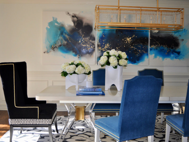 A modern dining room features a white table with a bouquet of white flowers, surrounded by black and blue chairs. A large abstract painting hangs on the wall, and a decorative light fixture is above.