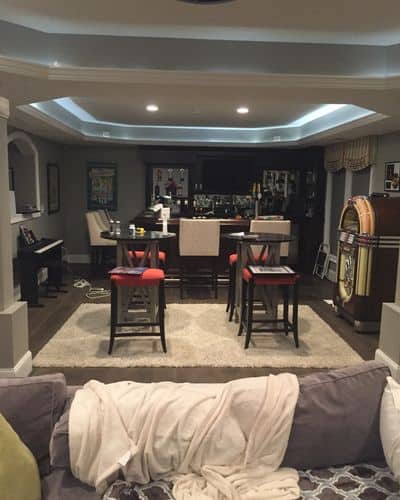 A home bar area with a high-top table and stools, a piano on the left, and a jukebox on the right. The ceiling has recessed lighting, and there's a sofa with a blanket in the foreground.