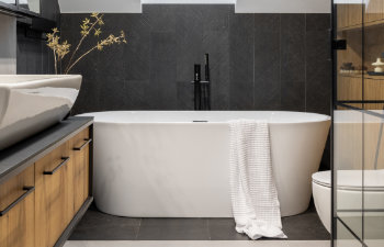 Modern bathroom featuring a freestanding white bathtub with a towel draped over it, black tile wall, wooden cabinetry, and a sink.