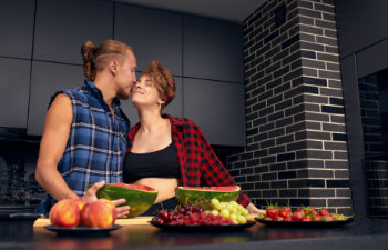 A couple in a kitchen, gently touching noses, with sliced watermelon, grapes, and apples on the counter.