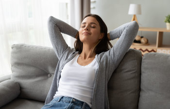 A woman is sitting on a gray sofa with her hands behind her head, eyes closed, and a peaceful expression. She is wearing a gray cardigan and a white top.